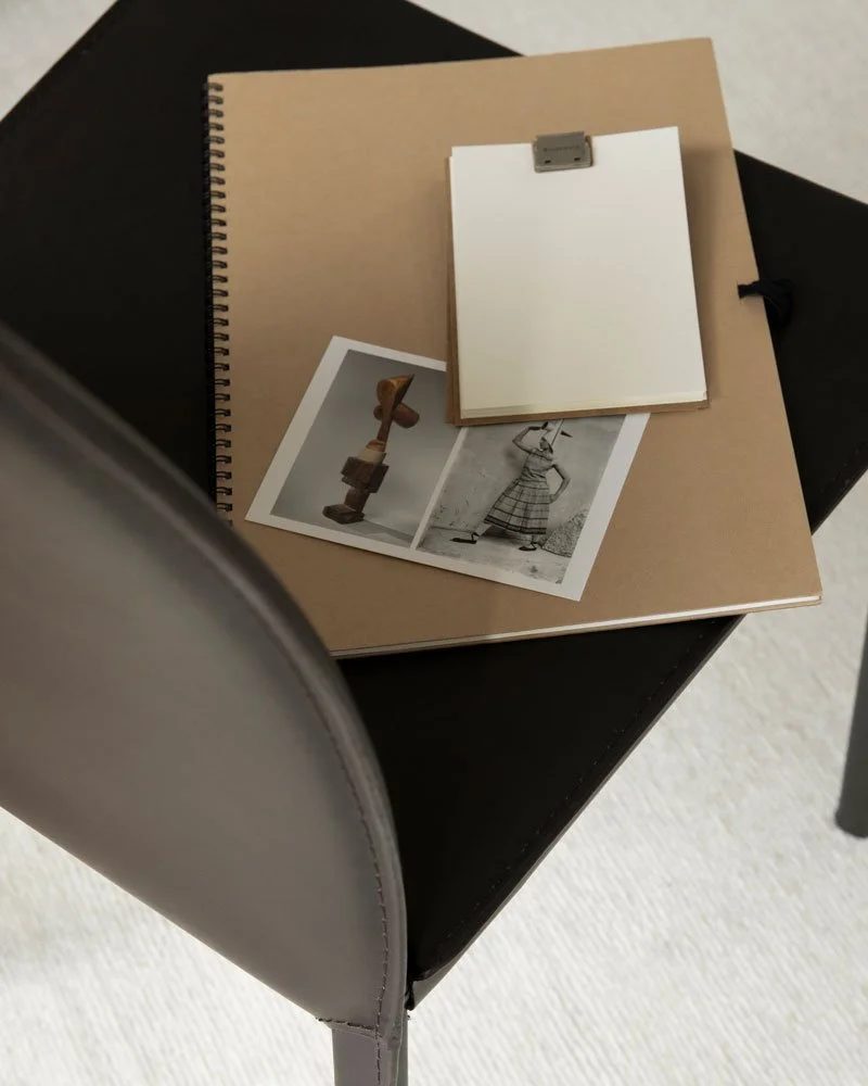 A black table with a beige notebook, a white notepad clipped with a metal clip, and two photographs of artwork and a woman in traditional clothing.