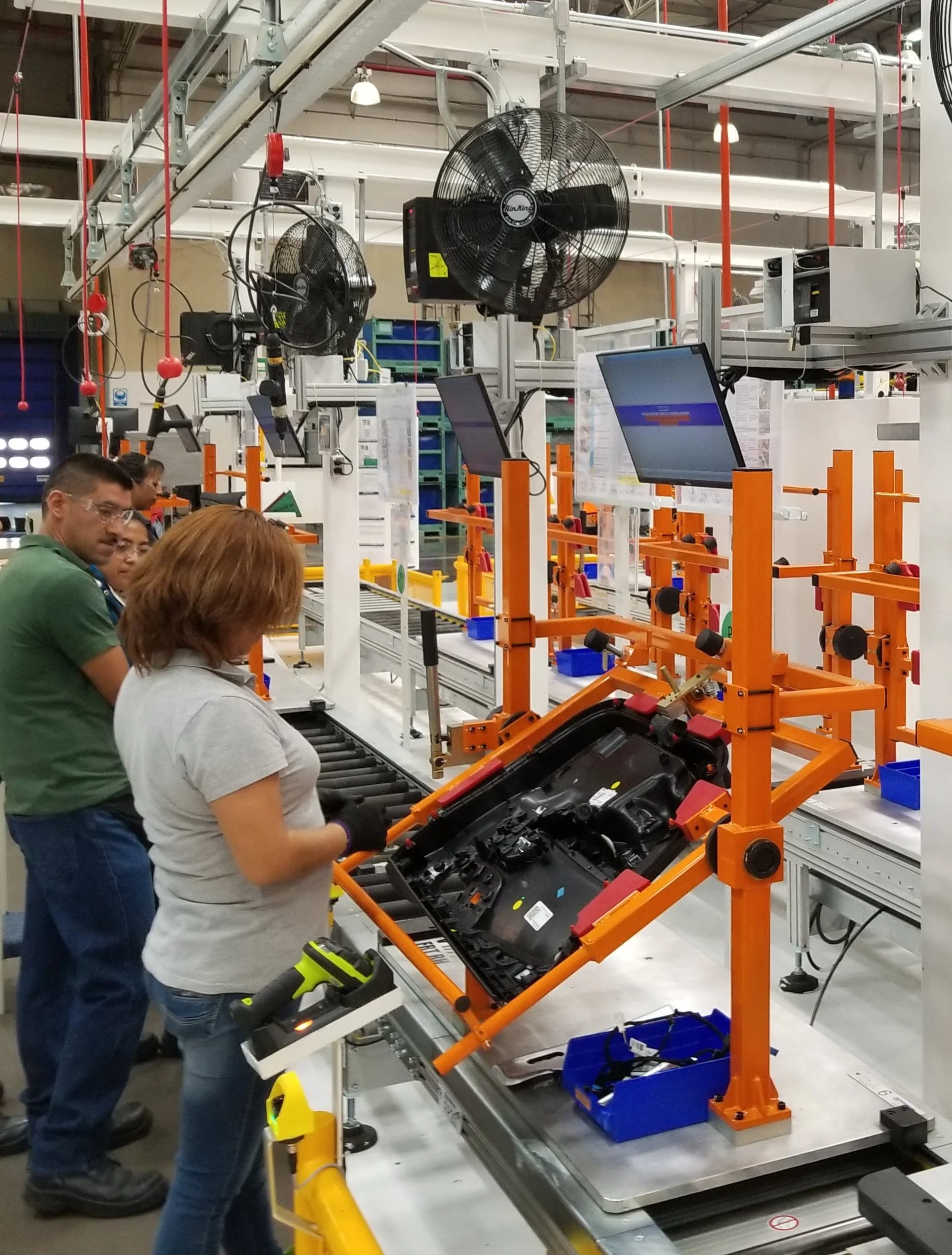Workers in a manufacturing facility assembling electronic devices on a conveyor belt with tools and equipment, surrounded by machinery and monitors.
