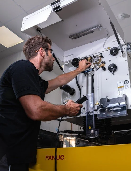 A man wearing safety glasses working on a large scientific or industrial machine, adjusting components with a tool, in an indoor laboratory or workshop.