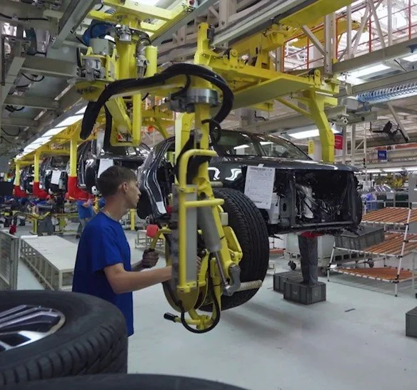 Automobile assembly line with a worker inspecting a partially assembled black car body, surrounded by robotic machinery in a factory setting.