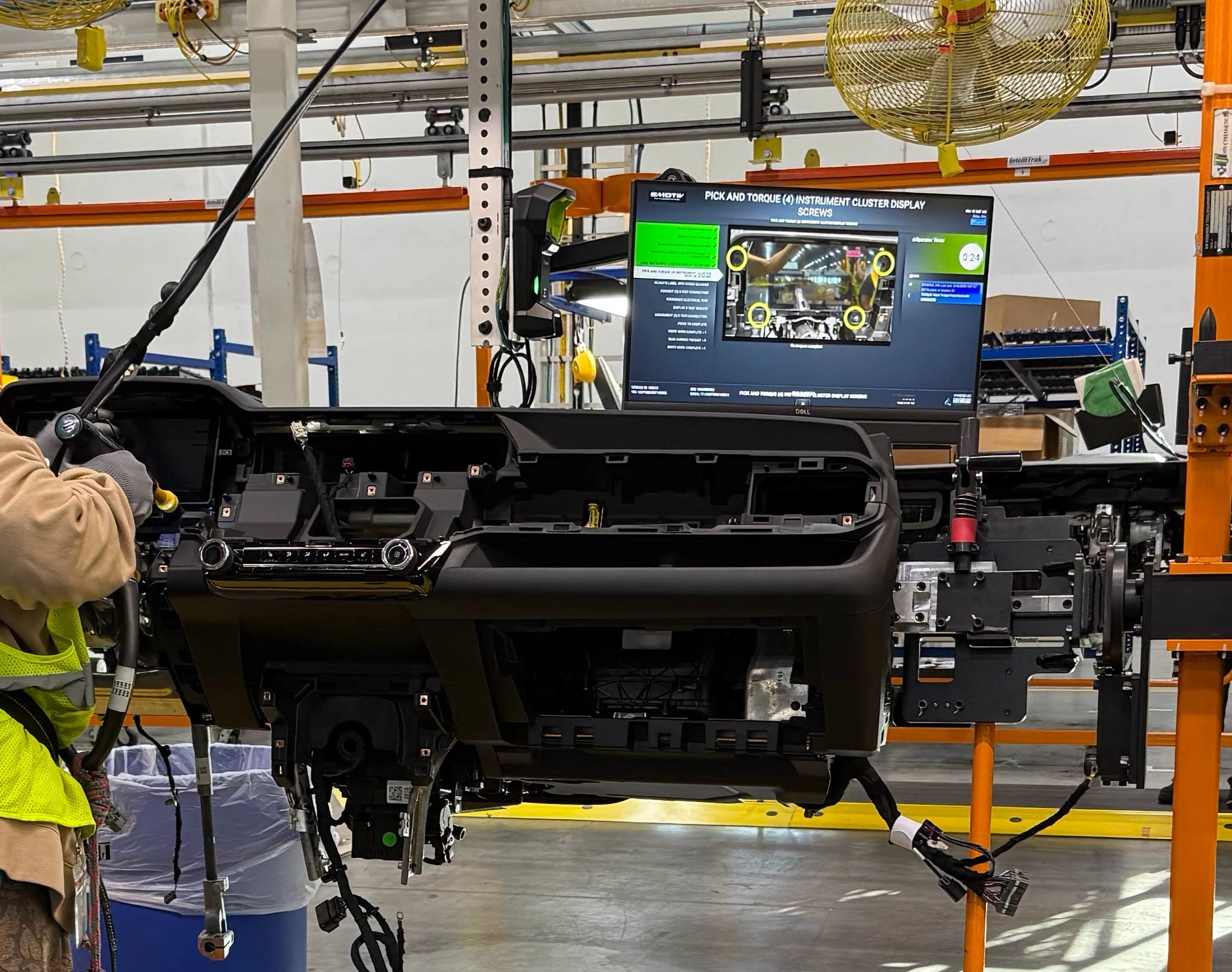 Inside a manufacturing facility, workers assemble automotive parts on an industrial assembly line, with a computer monitor displaying tools and instructions.