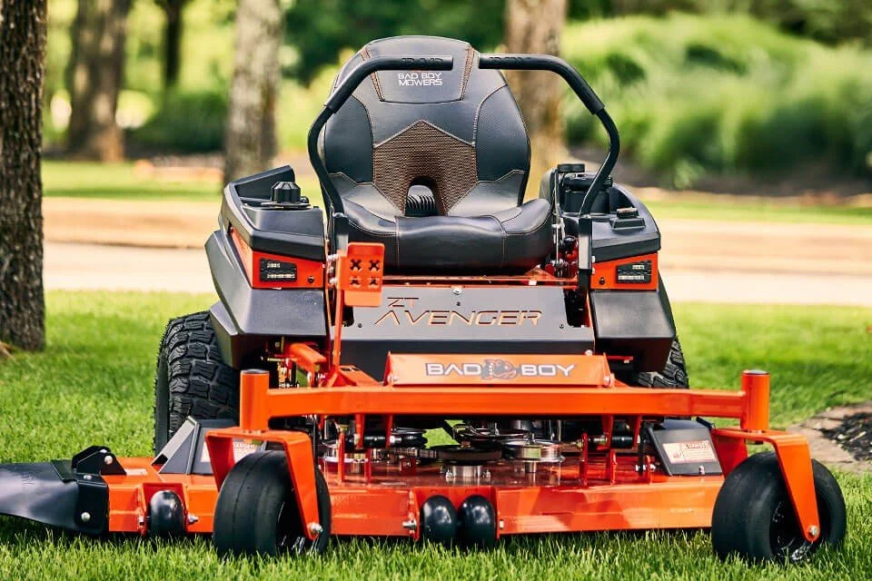 A zero-turn lawn mower with a black and orange color scheme, parked on a grass lawn with trees in the background.