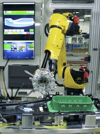 A yellow industrial robotic arm working on a manufacturing task inside a controlled environment, with a monitor displaying graphical data and controls, and a green tray on the workstation.