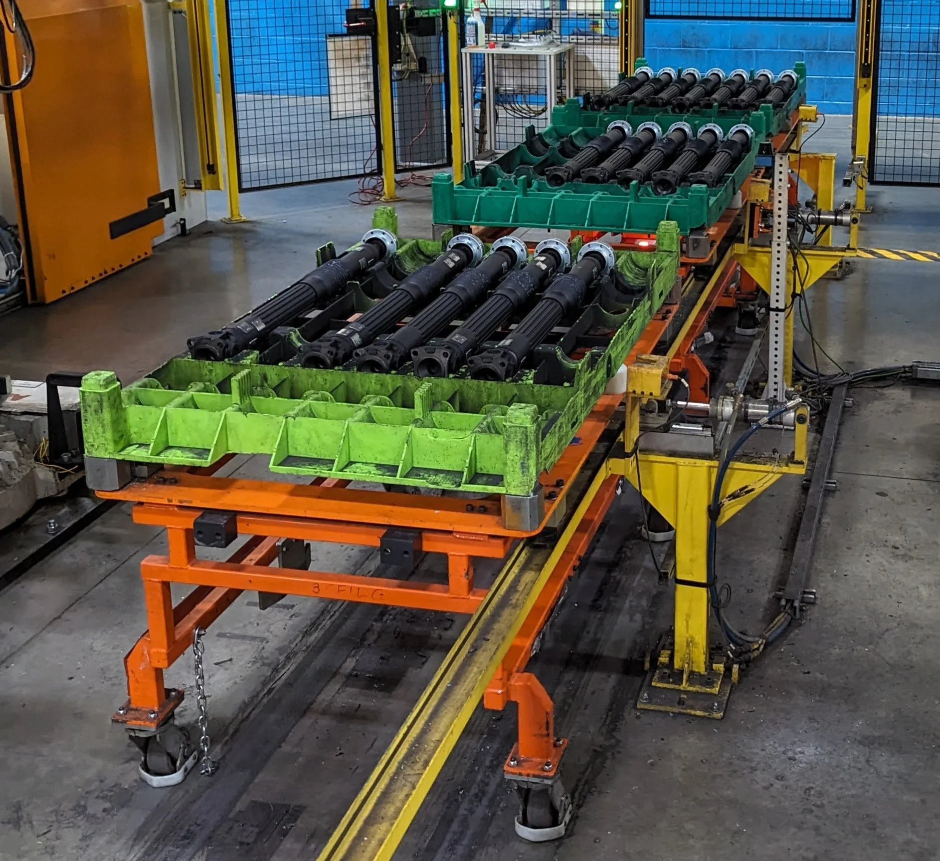 Industrial robotic manufacturing setup with long black cylindrical parts, green plastic trays, orange and yellow metal frames, and a blue wall in the background.