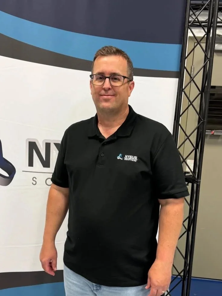 A man wearing glasses and a black polo shirt with the NYSUS Solutions logo standing in front of a banner with a partial logo and text, next to a metal framework.