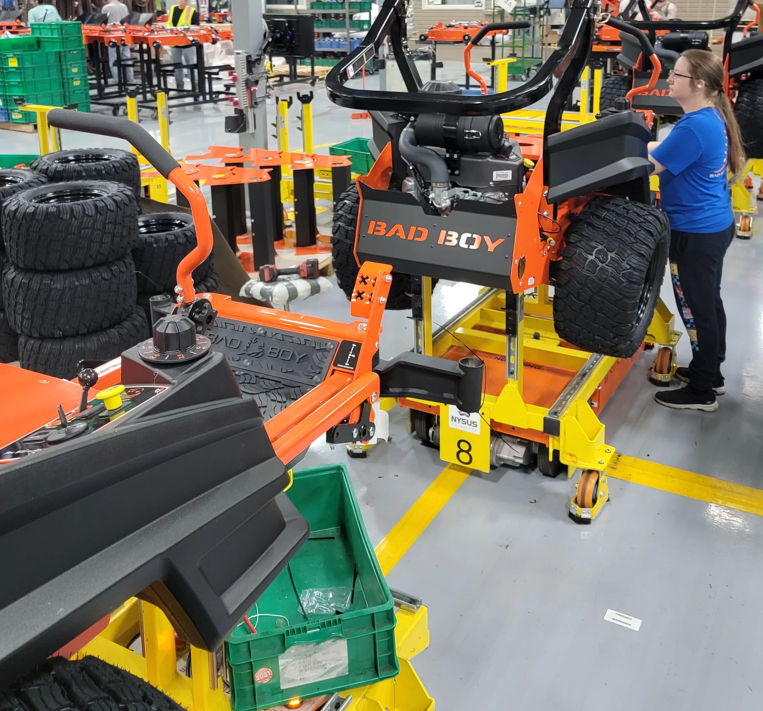 A person in a blue shirt operating a large orange and black machine labeled 'BAD BOY' in a warehouse or workshop setting with tires and green bins nearby.