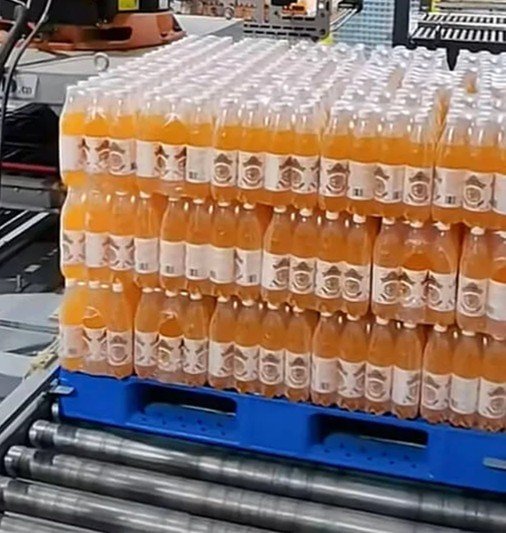 Bottles of orange-colored beverage in a factory setting, stacked on a blue pallet on a conveyor belt.