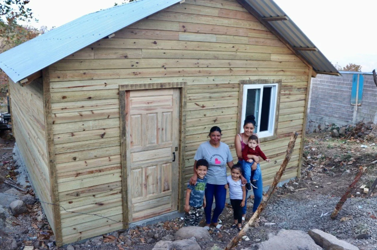 Photo of a group of four people, including women and children, standing outside a newly built small wooden house with a metal roof. The house has a wooden door and a window, and the surroundings show a construction site with rocks and debris.