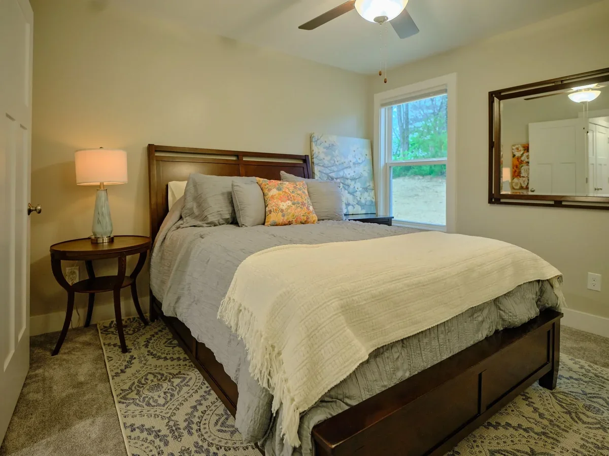 A cozy bedroom with a wooden bed frame, gray bedding, decorative pillows, a side table with a lamp, a window showing greenery outside, and a large mirror on the wall.