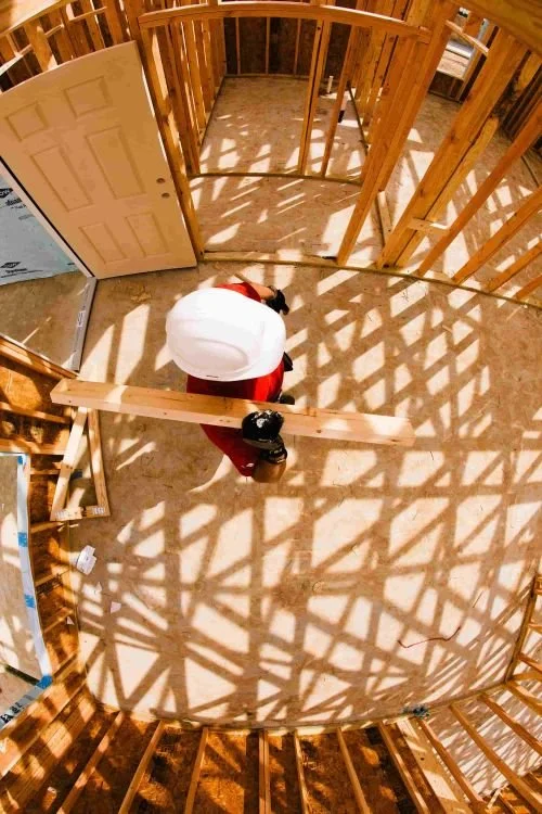 View from above of a construction site with wooden framing and a worker wearing a white hard hat and red shirt, working on a building structure in daylight.