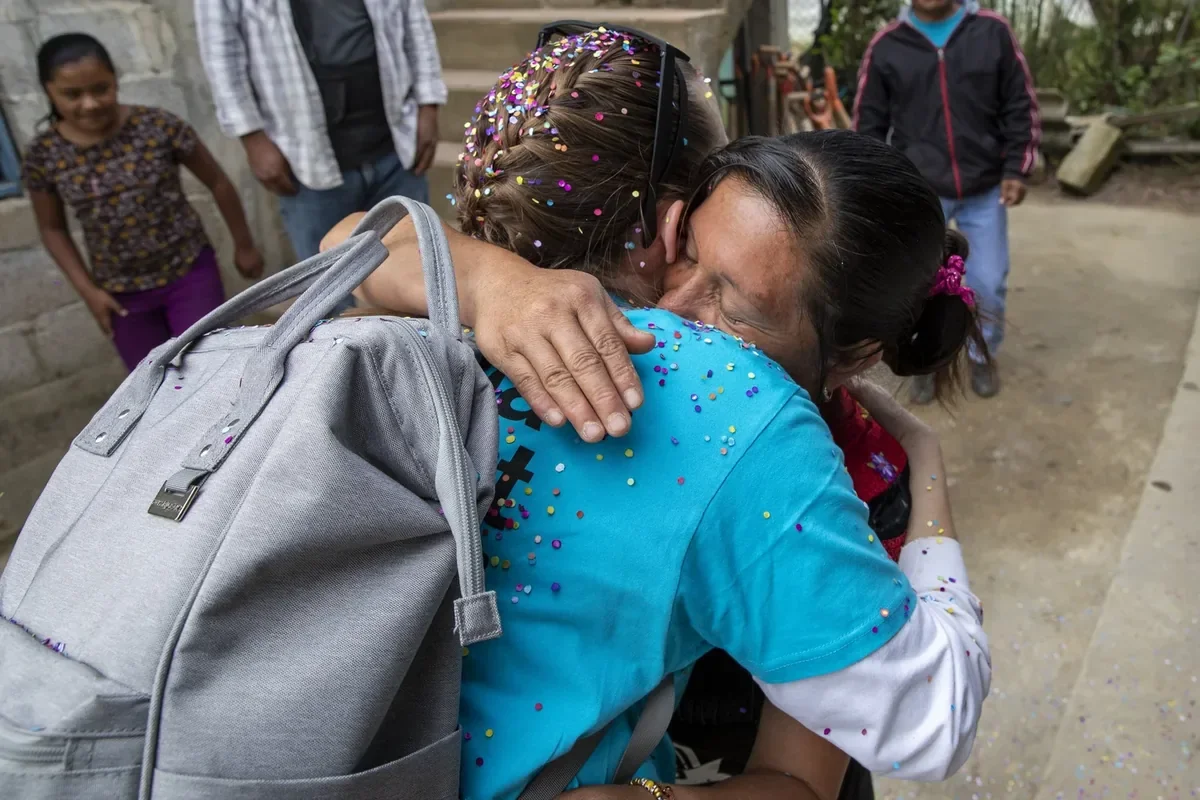 Two women hugging warmly, surrounded by a few children, with colorful confetti on their clothing and hair, outdoors.