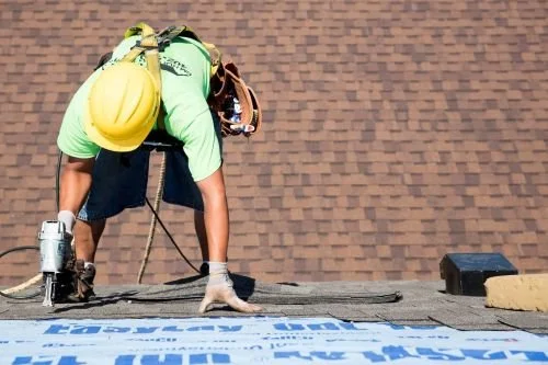 A construction worker wearing a yellow helmet, green shirt, and gloves, working on a roof with roofing tools. The worker is bent over, fixing shingles, with a brown bag tool pouch and safety harness, against a brick wall background.