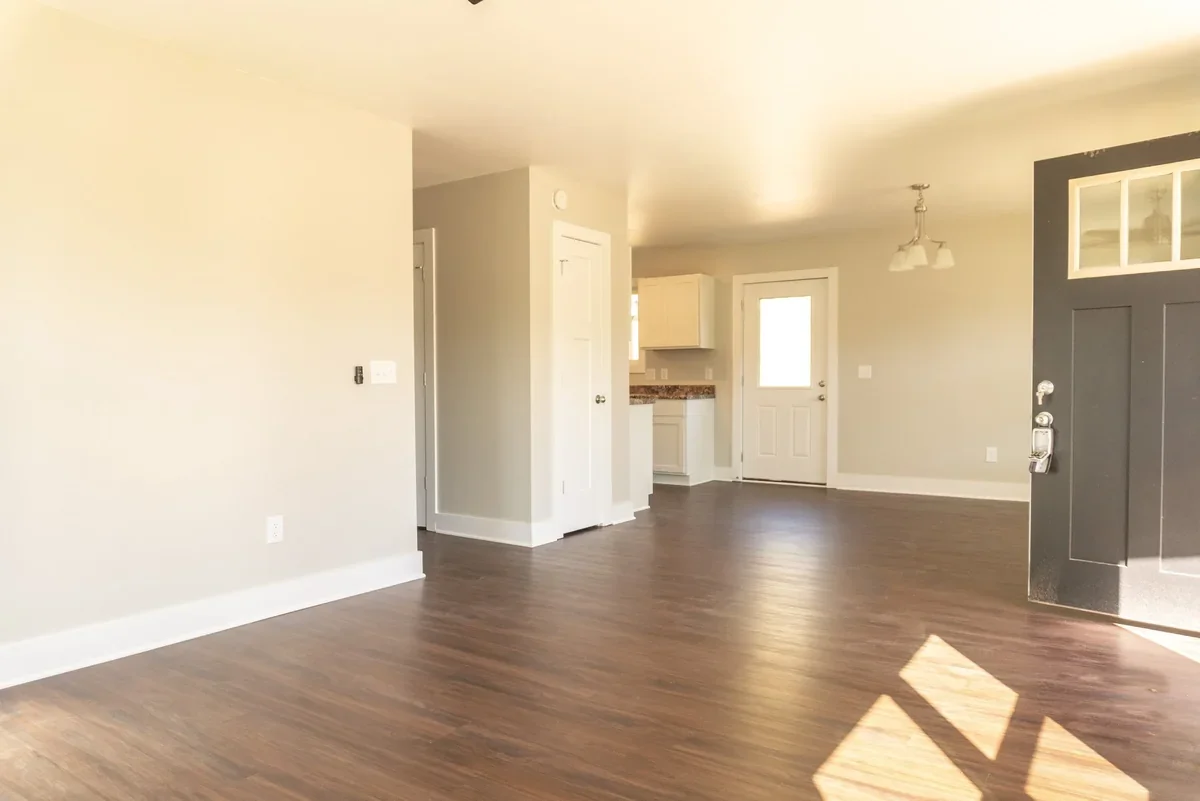 Empty living room with hardwood floors, beige walls, and white trim, leading to a doorway and a small kitchen area in the background.