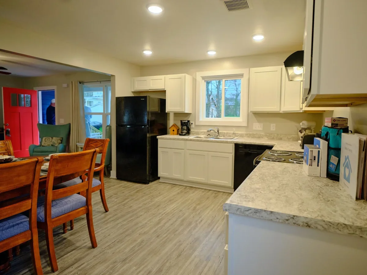 Kitchen and dining area with white cabinets, black refrigerator, stone countertops, and wooden dining table with chairs. Open doorway with a red door and a man outside visible in the background.