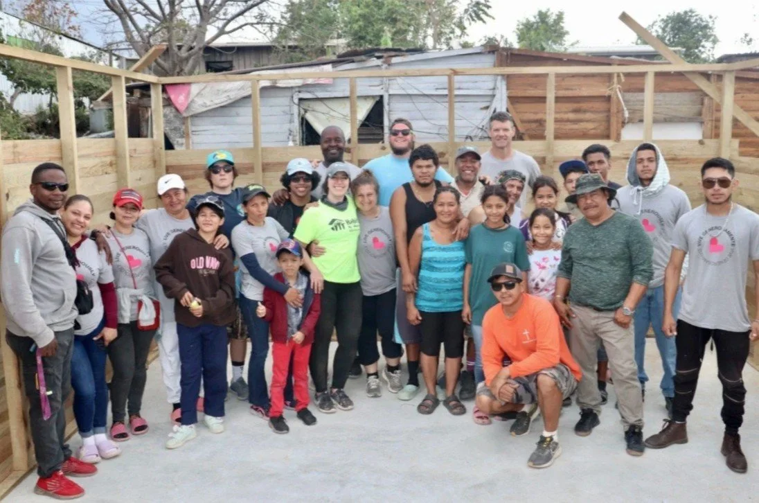 Group of diverse people standing together outdoors, some wearing casual clothes and hats, in front of a partially constructed wooden structure.