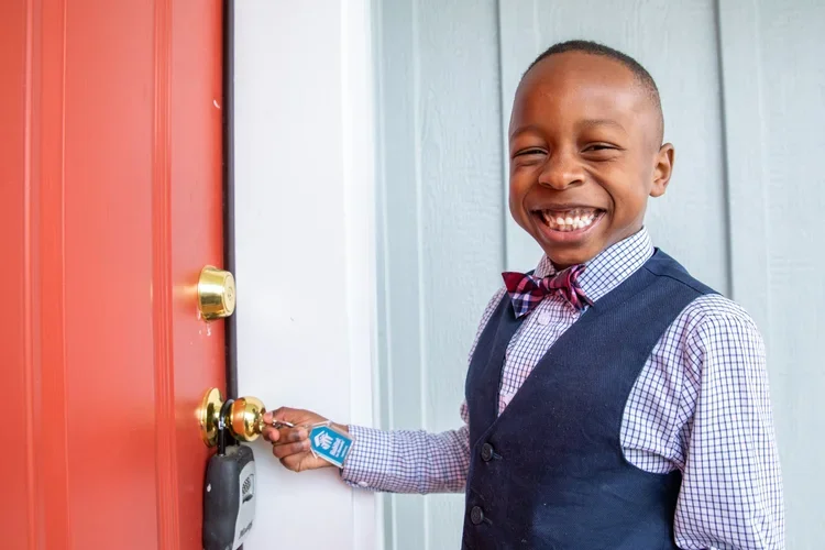 A young boy with a big smile, wearing a suit and bowtie, is standing at a door and holding a key to a lock with a keycard attached.