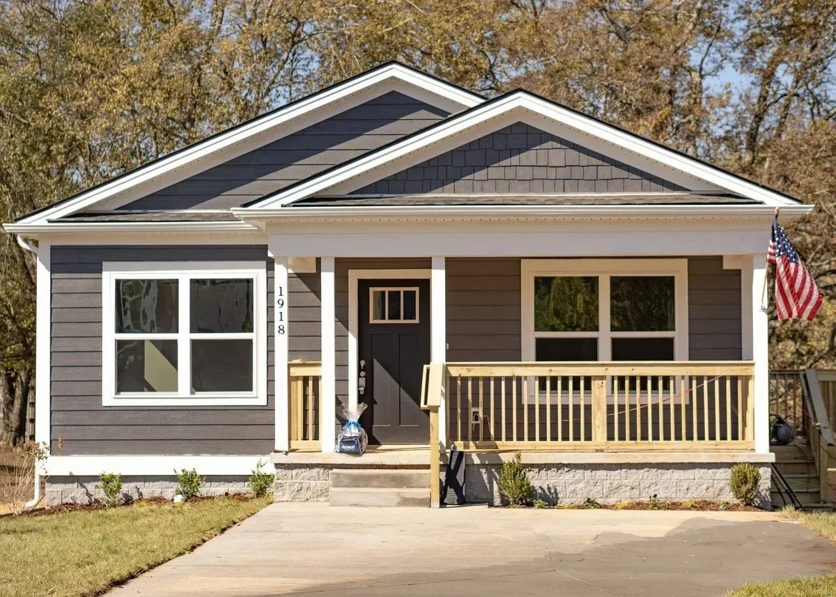 A newly constructed house with dark gray siding, white trim, a small front porch with a wooden railing, a front door, and two large windows. An American flag is on the right side, and a bag of supplies is near the front steps. The house number 1918 i
