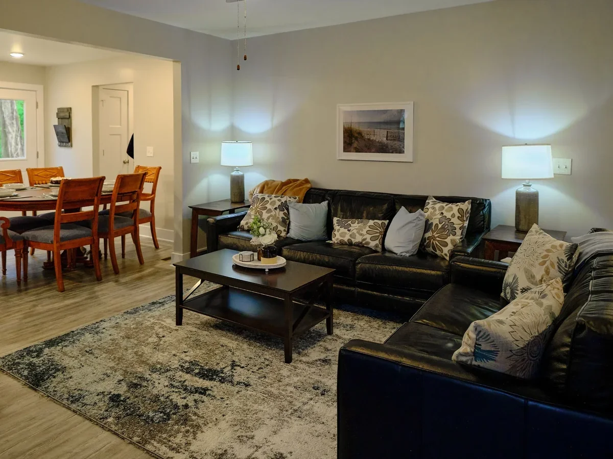 Living room with black leather sofas, patterned throw pillows, a wooden coffee table with a tray and decorative items, lamps on side tables, a framed beach picture on the wall, and a dining area with wooden chairs and a table in the background.