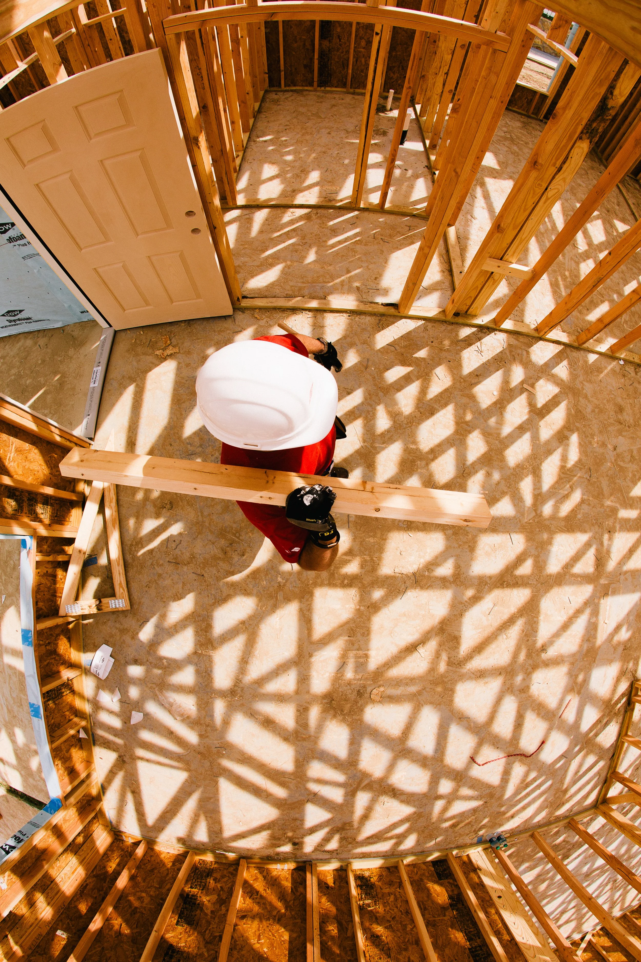 View from above of a construction site with wooden framing and a worker wearing a white hard hat and red shirt, working on a building structure in daylight.