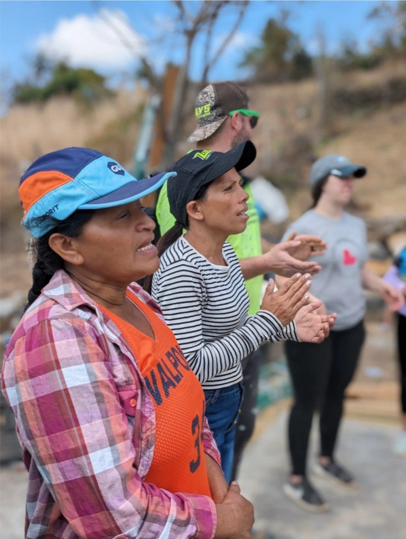 A group of people standing outdoors, some with their hands clasped together and eyes closed, appearing to be praying or in reflection. They are dressed casually and wearing hats and sunglasses, with a natural background of trees and a blue sky.