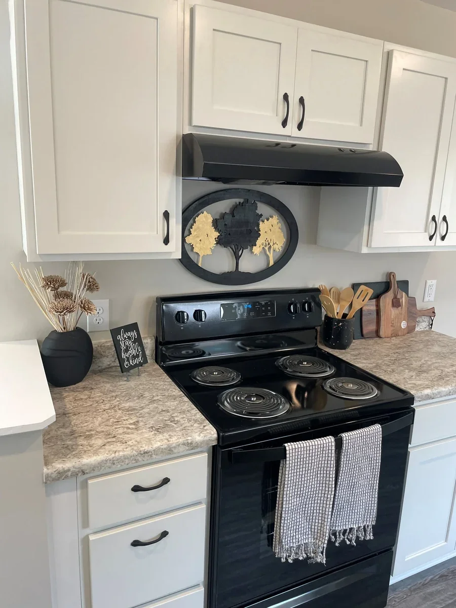 A kitchen featuring white cabinets, a black stove with two towels hanging from the handle, and a granite countertop decorated with a black vase of dried flowers, a small black sign, wooden utensils, and cutting boards. There is also a black range hoo