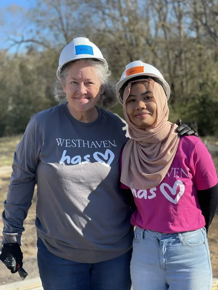 Two women standing outdoors wearing construction helmets and grey shirts with the message 'Westhaven has ❤️'. The woman on the left has grey hair and is holding gloves, while the woman on the right is wearing a beige hijab and a pink shirt. Both are smiling and looking at the camera, with trees and clear sky in the background.