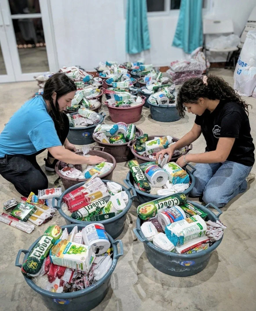 Two women sorting through baskets of canned and packaged food in a room with blue curtains.
