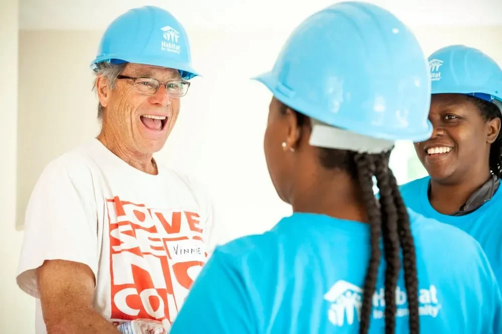 Three people wearing blue Habitat for Humanity helmets and shirts, smiling and talking indoors.