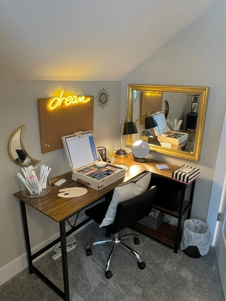 Home office workspace with a wooden desk, black chair, mirror, gold frame, black and gold desk lamps, neon yellow 'dream' sign on wall, moon and sun wall decor, organized art supplies, and a trash bin.