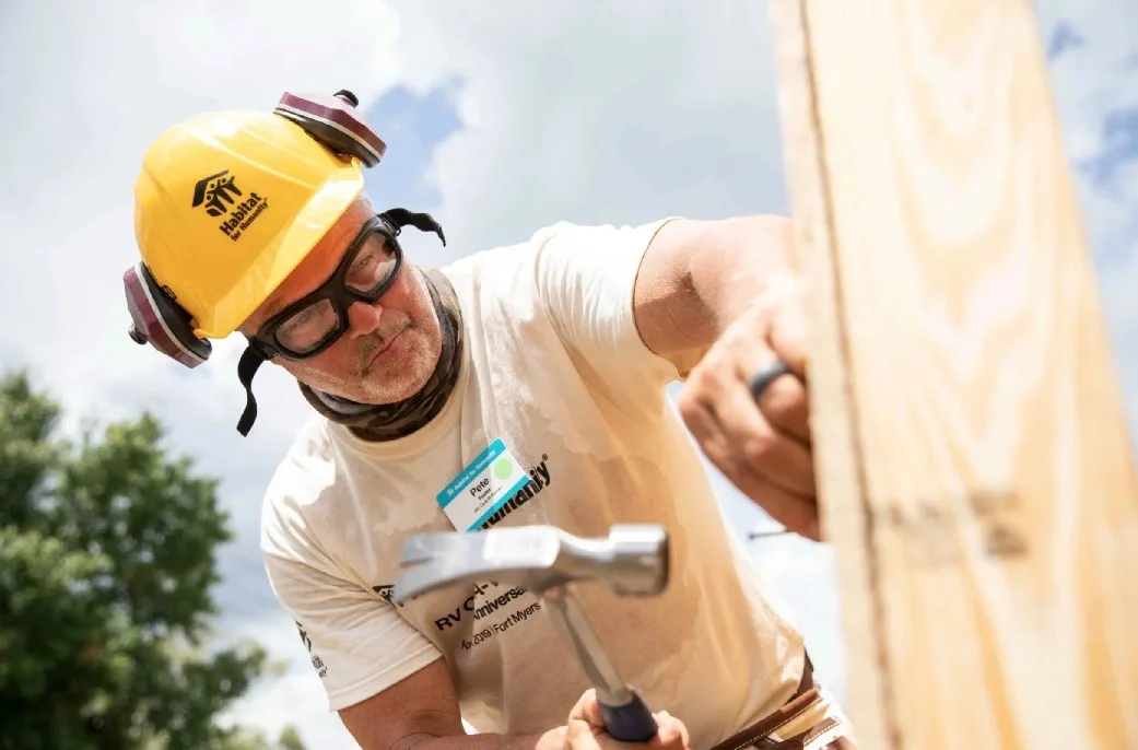 A man wearing safety glasses, a yellow helmet with the Habitat for Humanity logo, and a T-shirt with a Habitat for Humanity logo, is hammering a nail into a wooden structure outdoors on a sunny day.