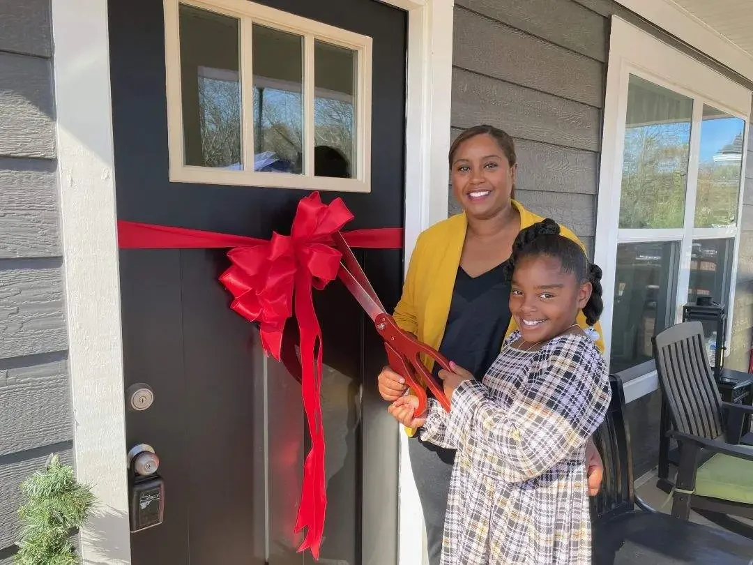 A woman and girl are standing outside a house beside a front door decorated with a large red bow and ribbon, holding scissors for a ribbon-cutting ceremony, both smiling.