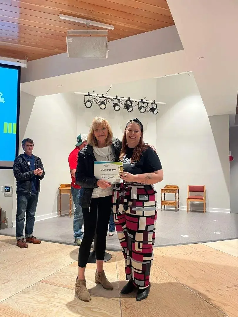 Two women standing on a stage holding a certificate and a trophy, with three people in the background. One woman is wearing a black jacket and beige shoes, and the other is dressed in a patterned jumpsuit. The setting appears to be an indoor event or ceremony.