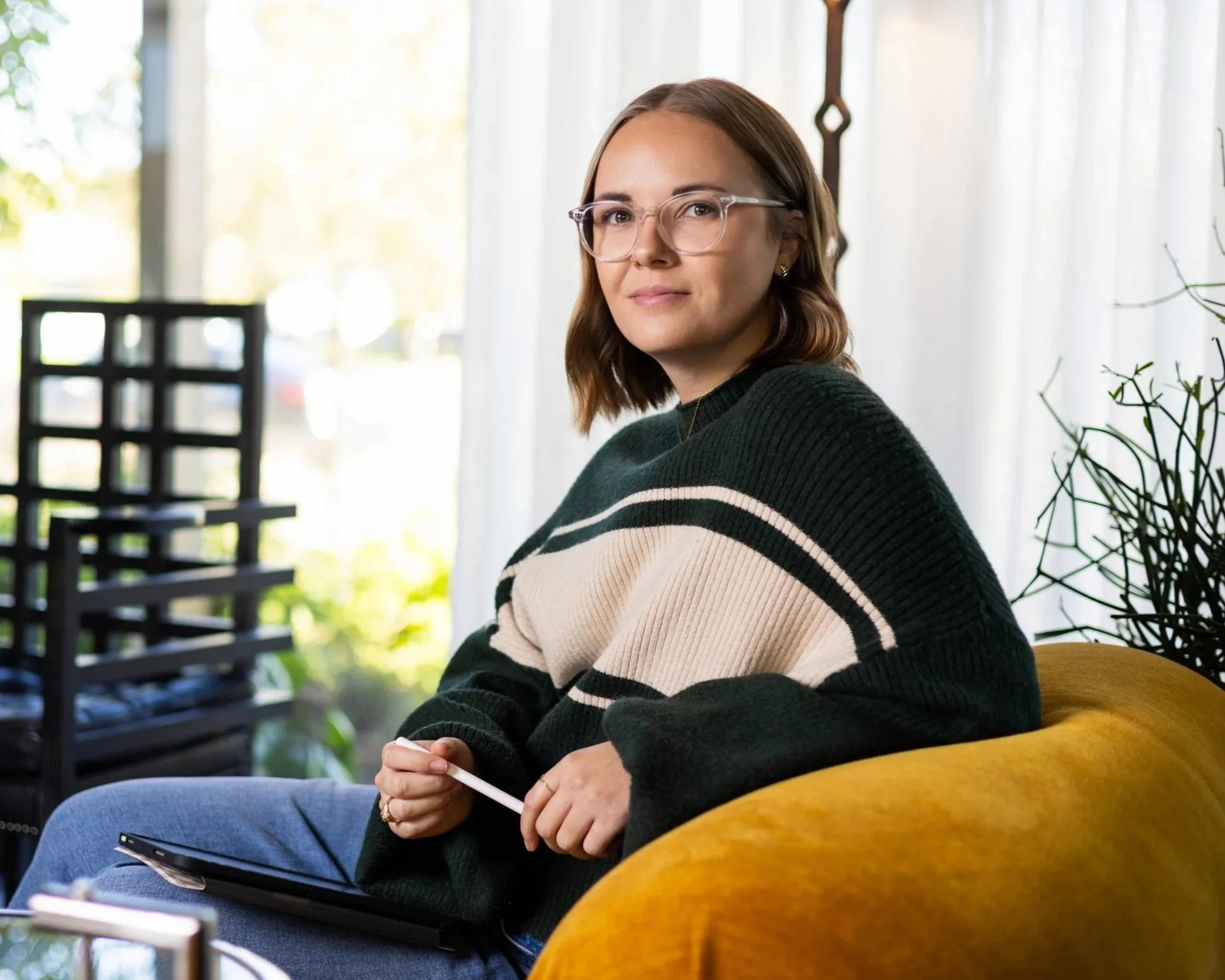 Young woman with shoulder-length brown hair, glasses, and a black and cream striped sweater sitting on a yellow couch with a tablet and stylus in her hands, in a well-lit room with plants and large windows.