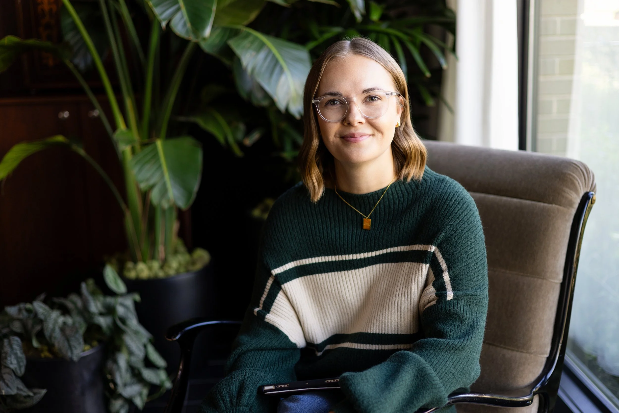 A young woman with glasses and blonde hair sitting in a chair near large green plants and a window, smiling at the camera.