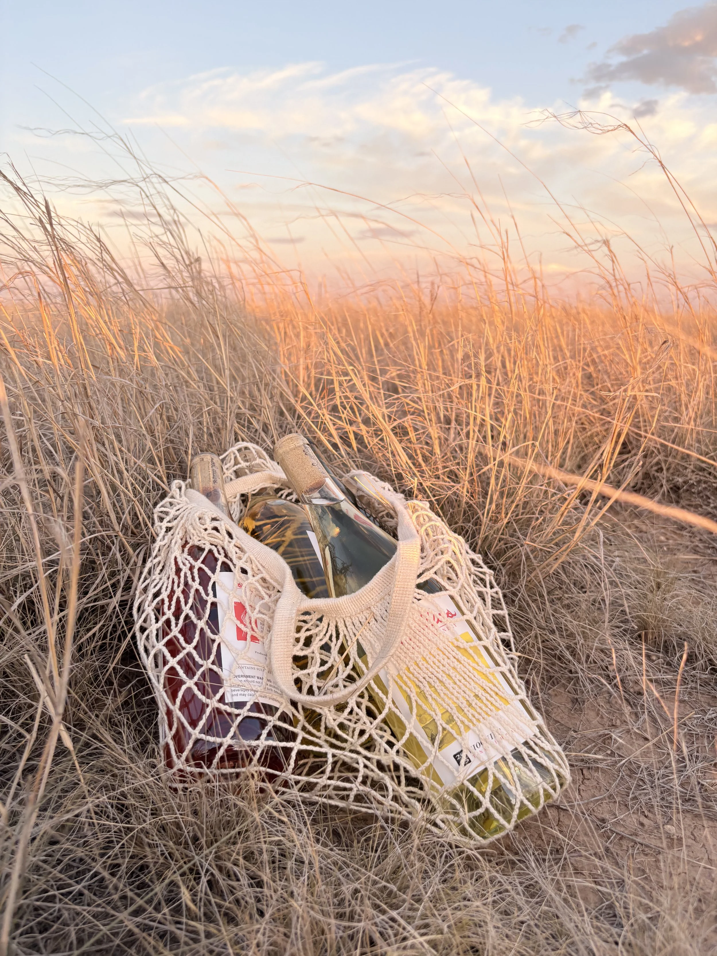 Glass bottles of Sod Cellars wine inside a netted cloth bag on a prairie field during sunset.
