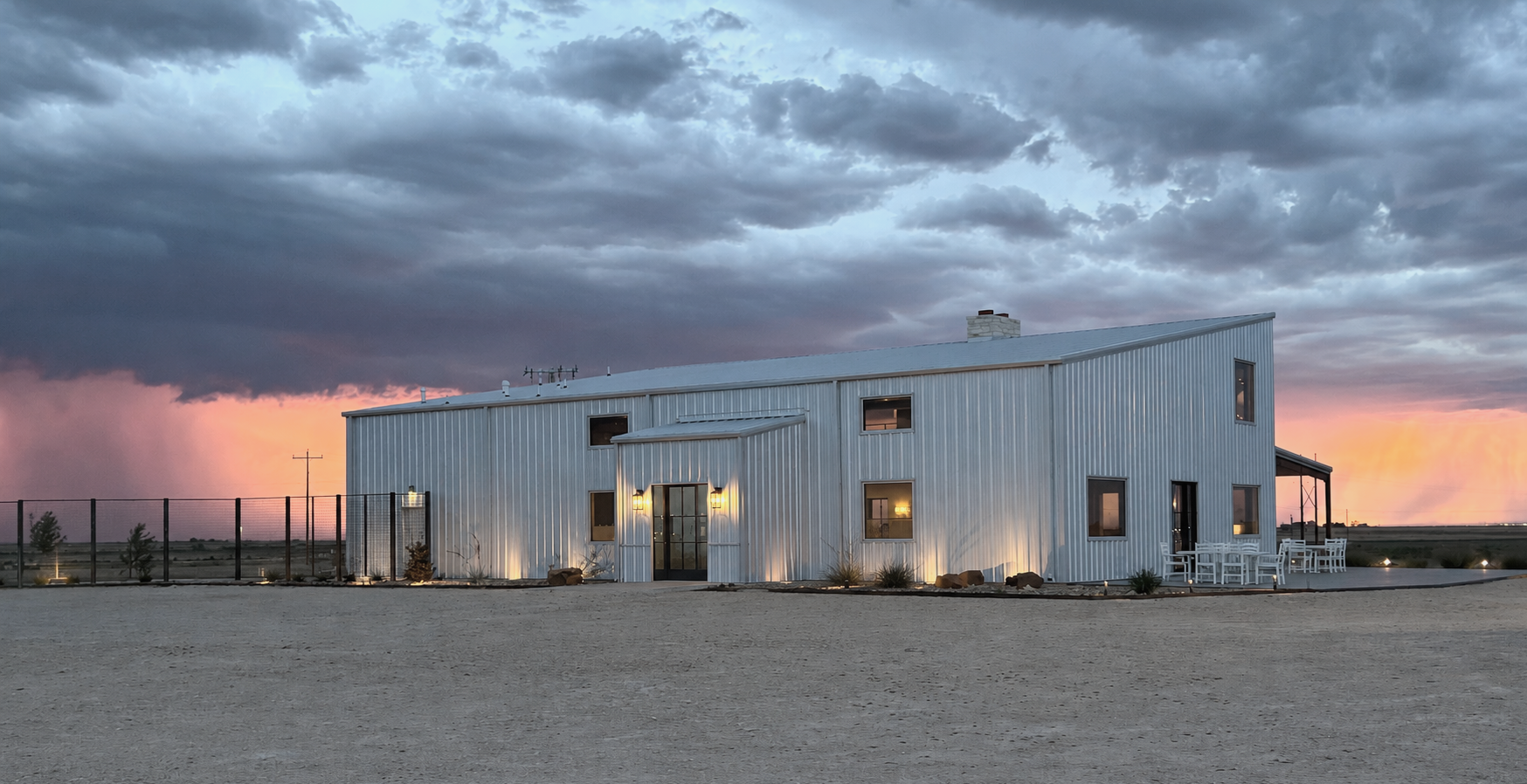 Modern metal building with large windows and outdoor seating, set against a dramatic sunset sky with dark clouds, in a rural landscape.