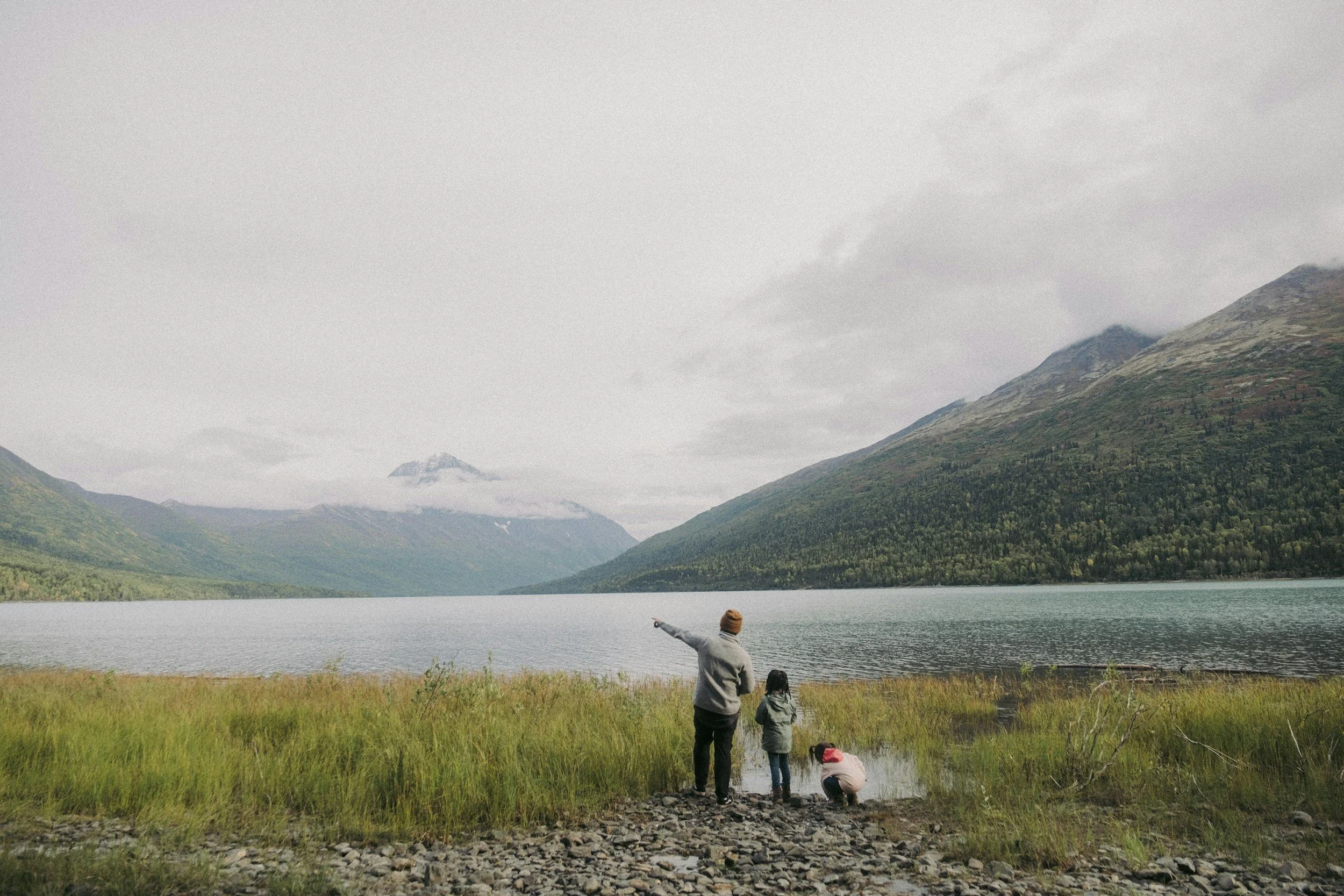 Three people, two children and an adult, enjoying a day near a lake in a mountainous area, with one of the adults pointing towards the distance.