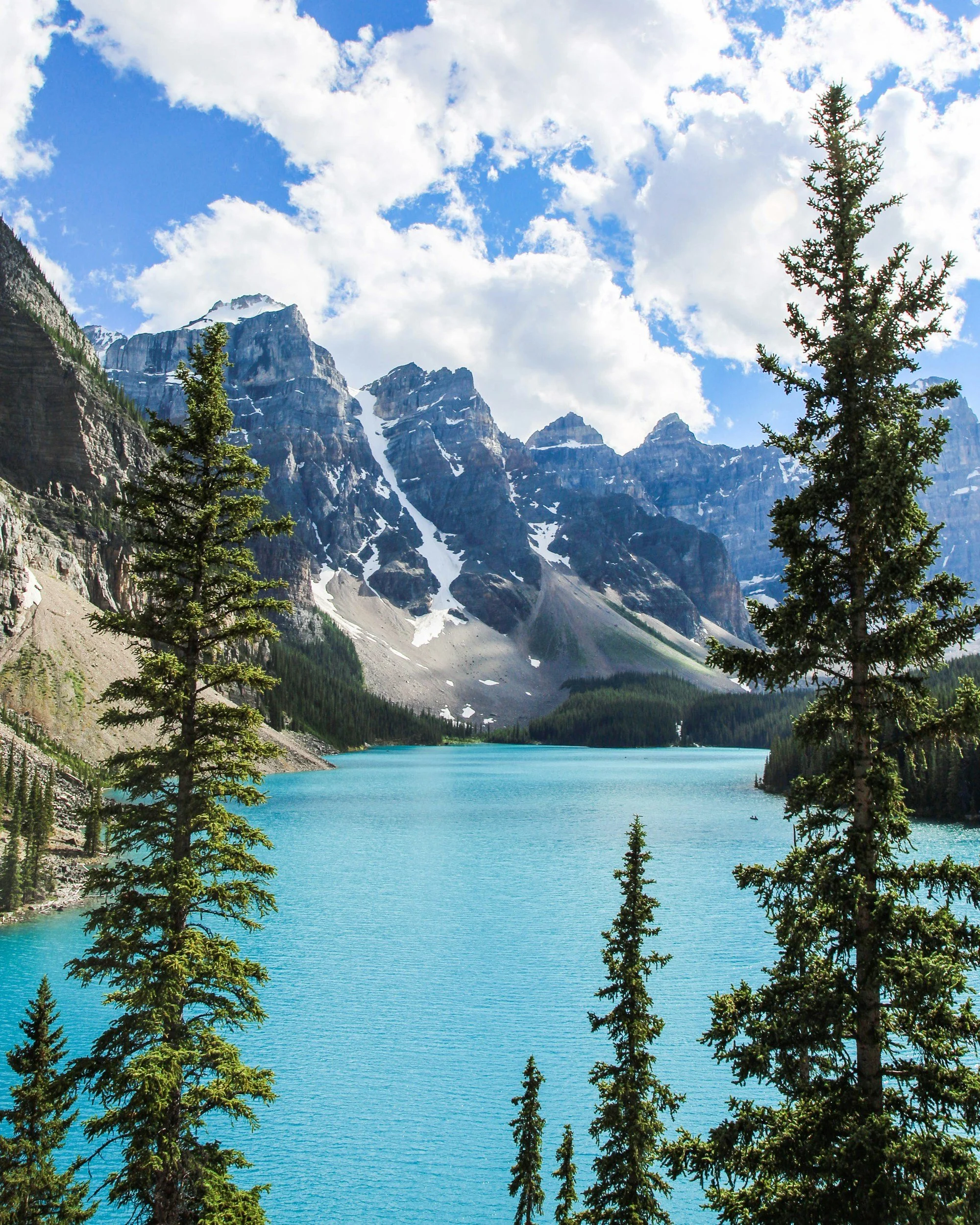 A mountain lake with turquoise water surrounded by pine trees and snow-capped peaks under a partly cloudy sky.