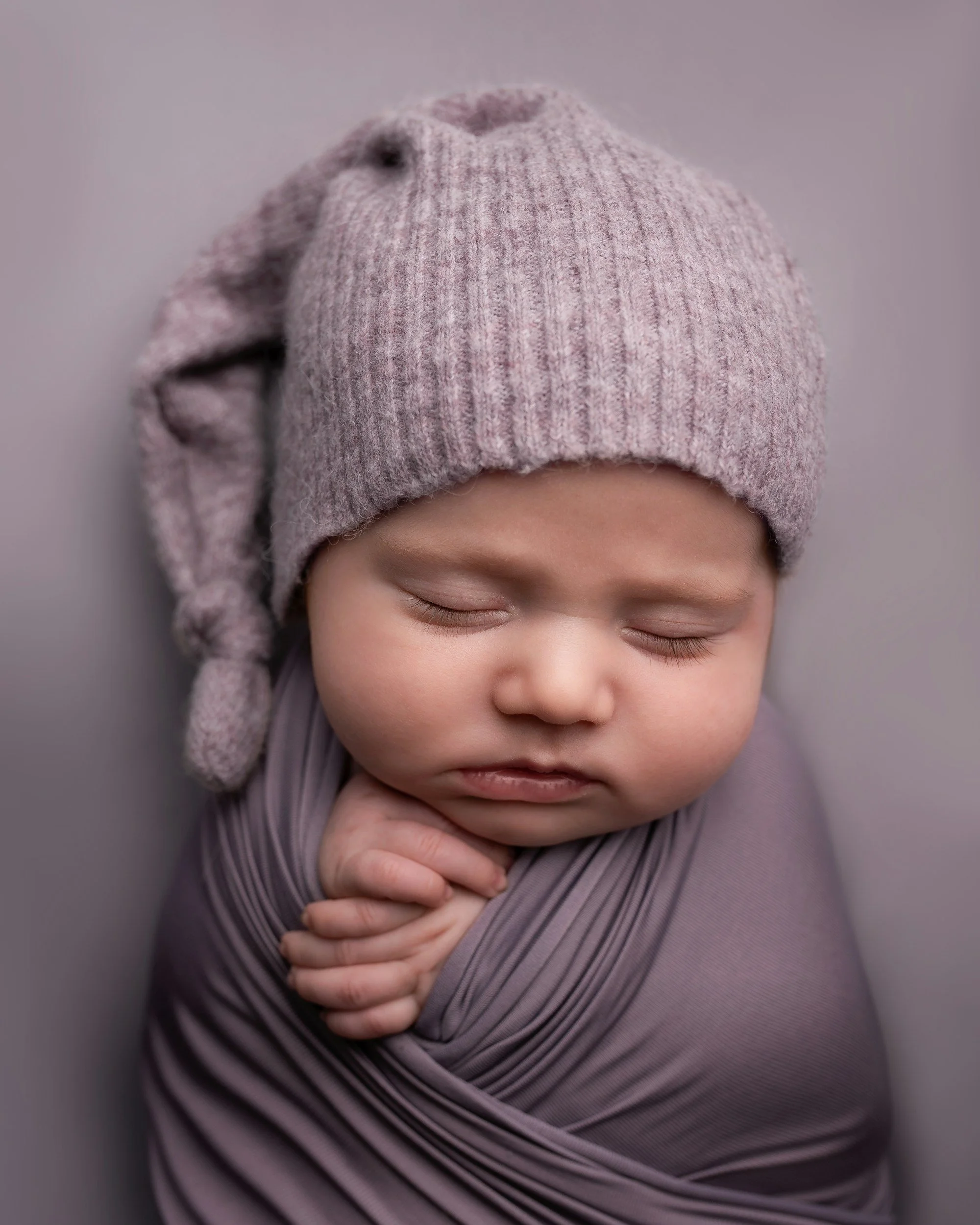 A close-up of a sleeping baby wearing a pink knit hat and wrapped in a taupe blanket.