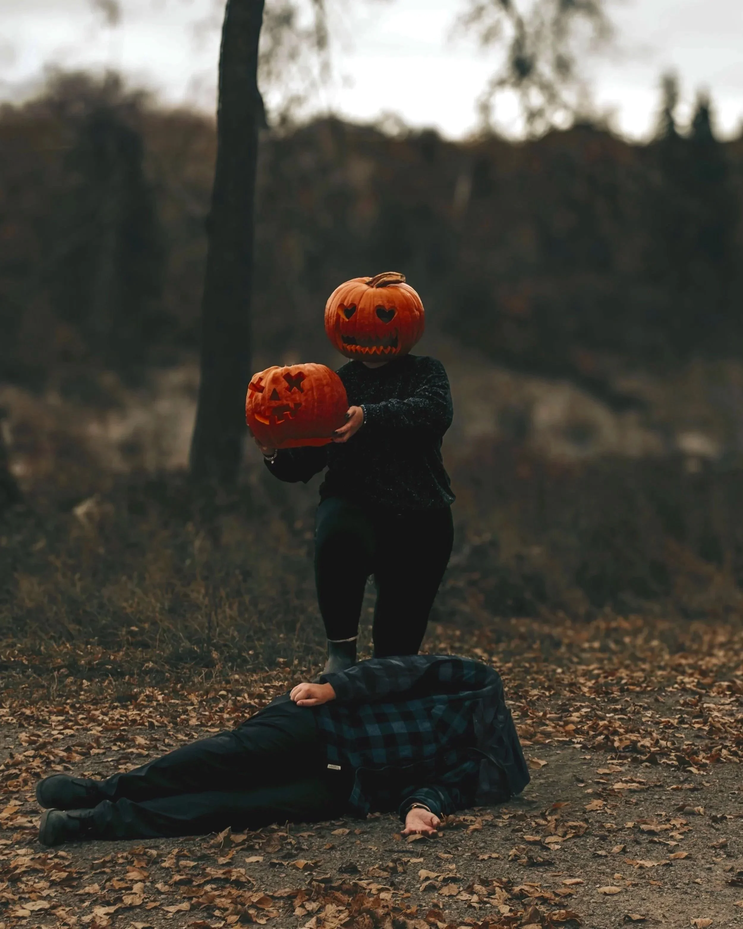 Person standing outdoors holding two carved Halloween pumpkins with jack-o'-lantern faces, while another person is lying on the ground with their hand touching their head, surrounded by fallen leaves.