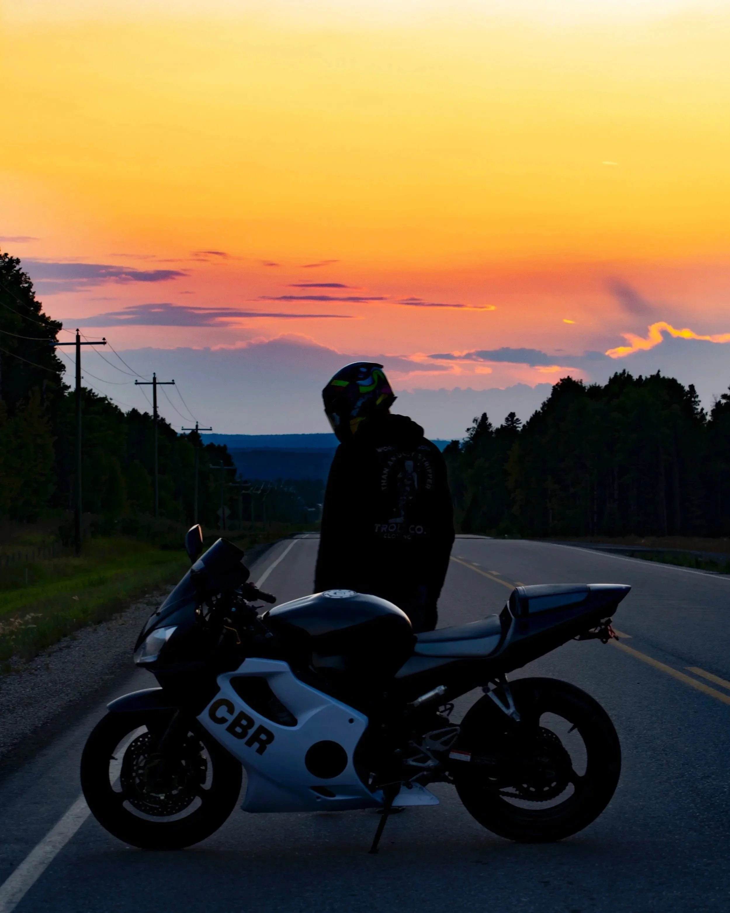 A person wearing a helmet and black jacket standing next to a black and white sport motorcycle on an empty road during sunset, with a colorful sky and trees on either side.
