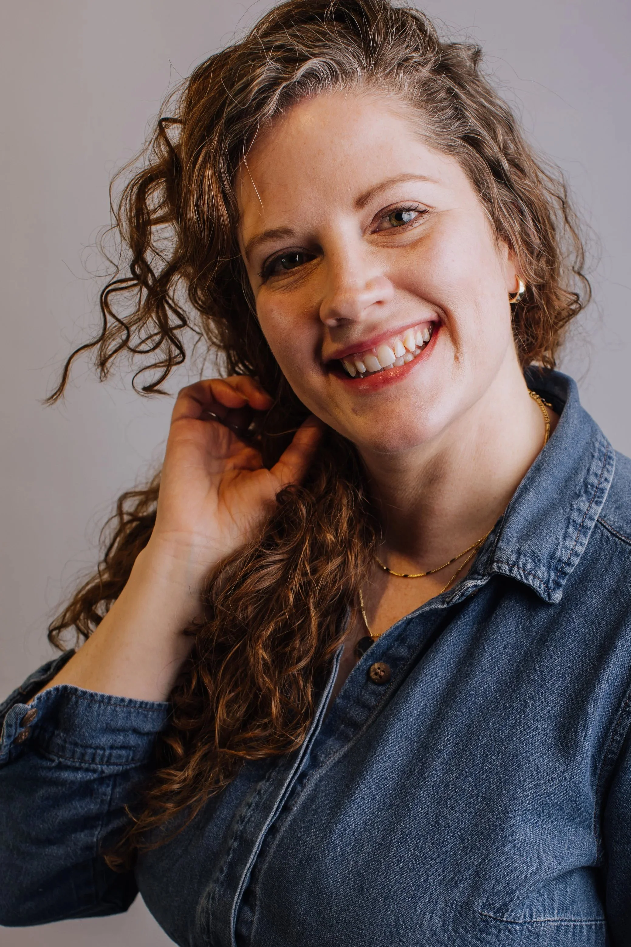 A woman with long, curly brown hair, smiling and touching her hair, wearing a denim shirt and gold jewelry, against a plain gray background.