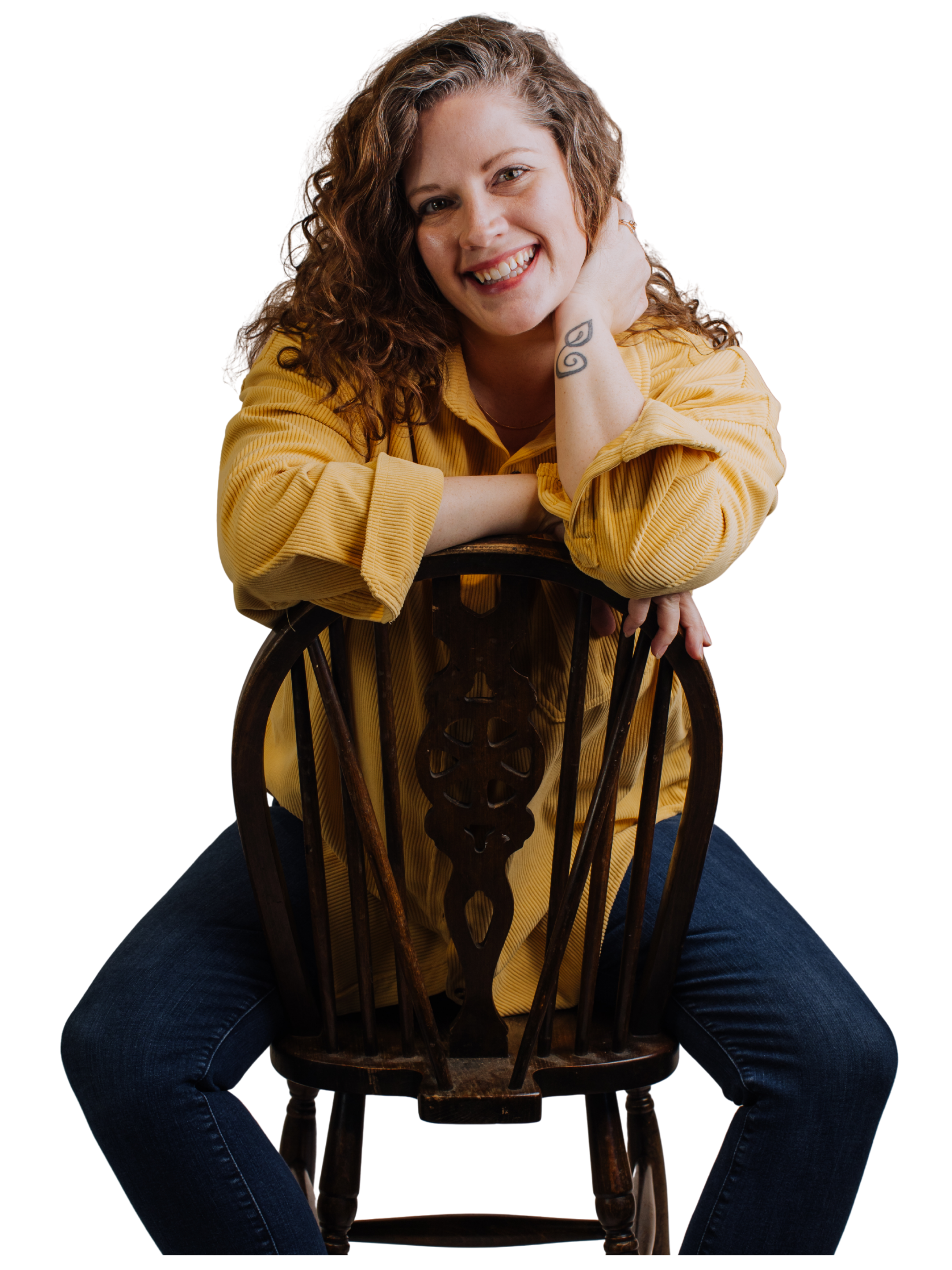 A woman with curly hair smiling, sitting on the floor with legs apart, leaning on a wooden chair, wearing a yellow shirt and jeans.