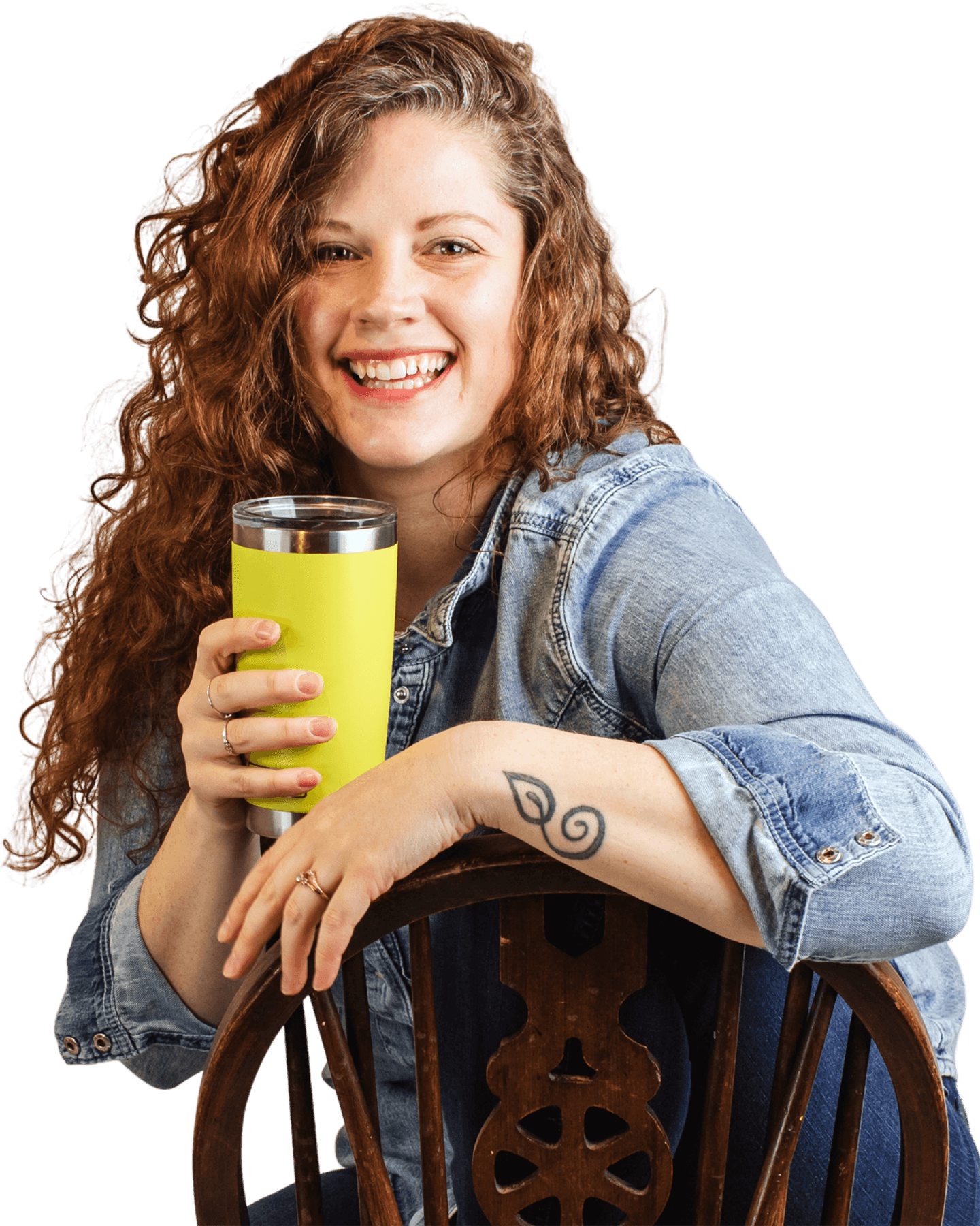 A woman with curly red hair smiling and holding a yellow travel mug. She is sitting in a wooden chair, wearing a denim jacket with rolled-up sleeves, and has a spiral tattoo on her forearm.