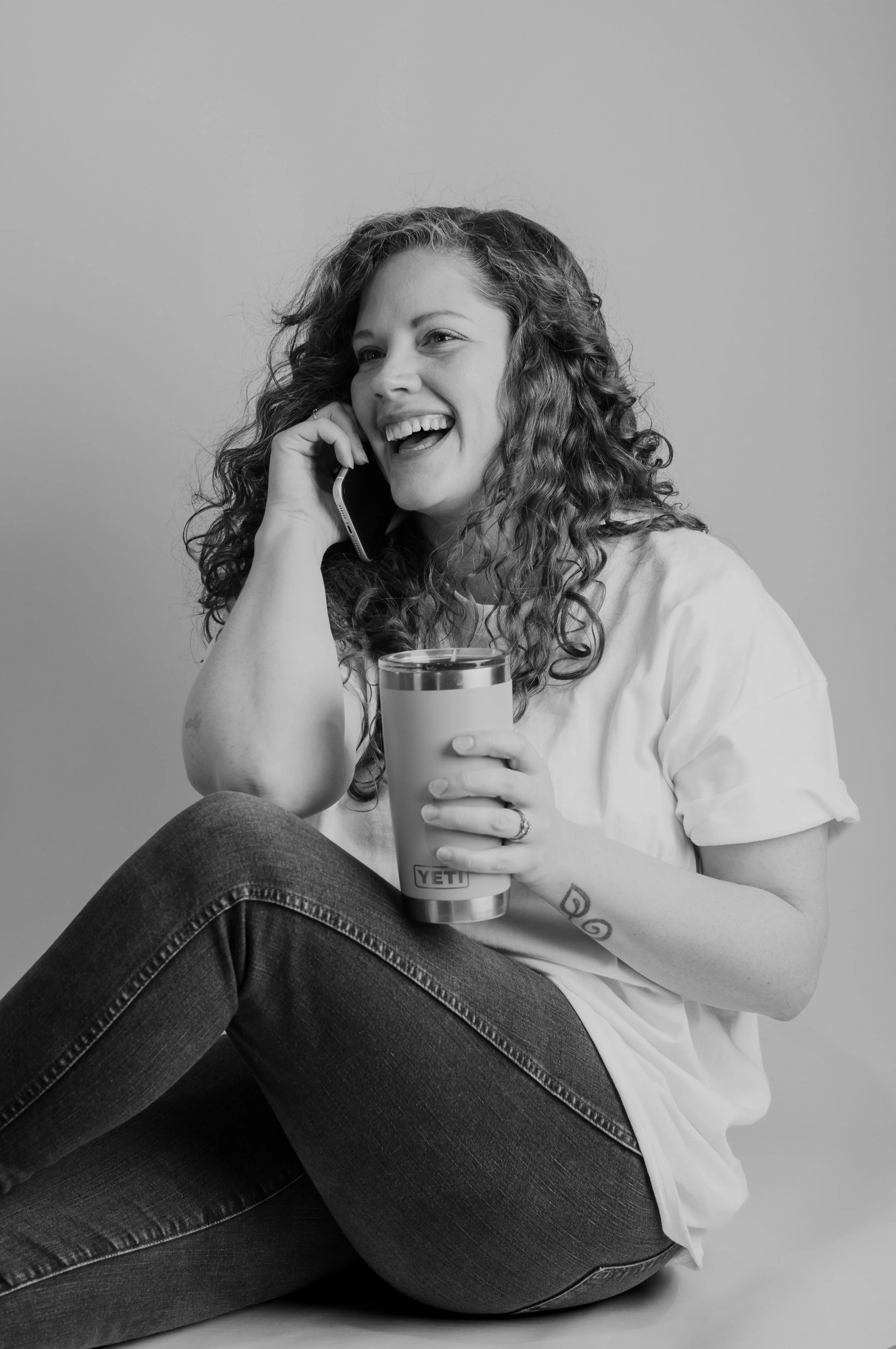 Black and white photo of a woman with curly hair smiling, talking on a phone, holding a tumbler, sitting with one leg crossed. She has tattoos on her arm and is wearing a white shirt and dark jeans.