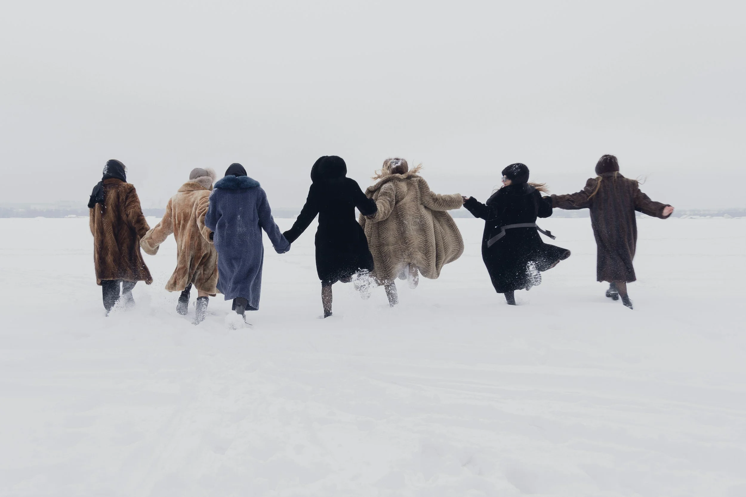 group of 7 people in heavy winter coats holding hands and running away from the camera through snow