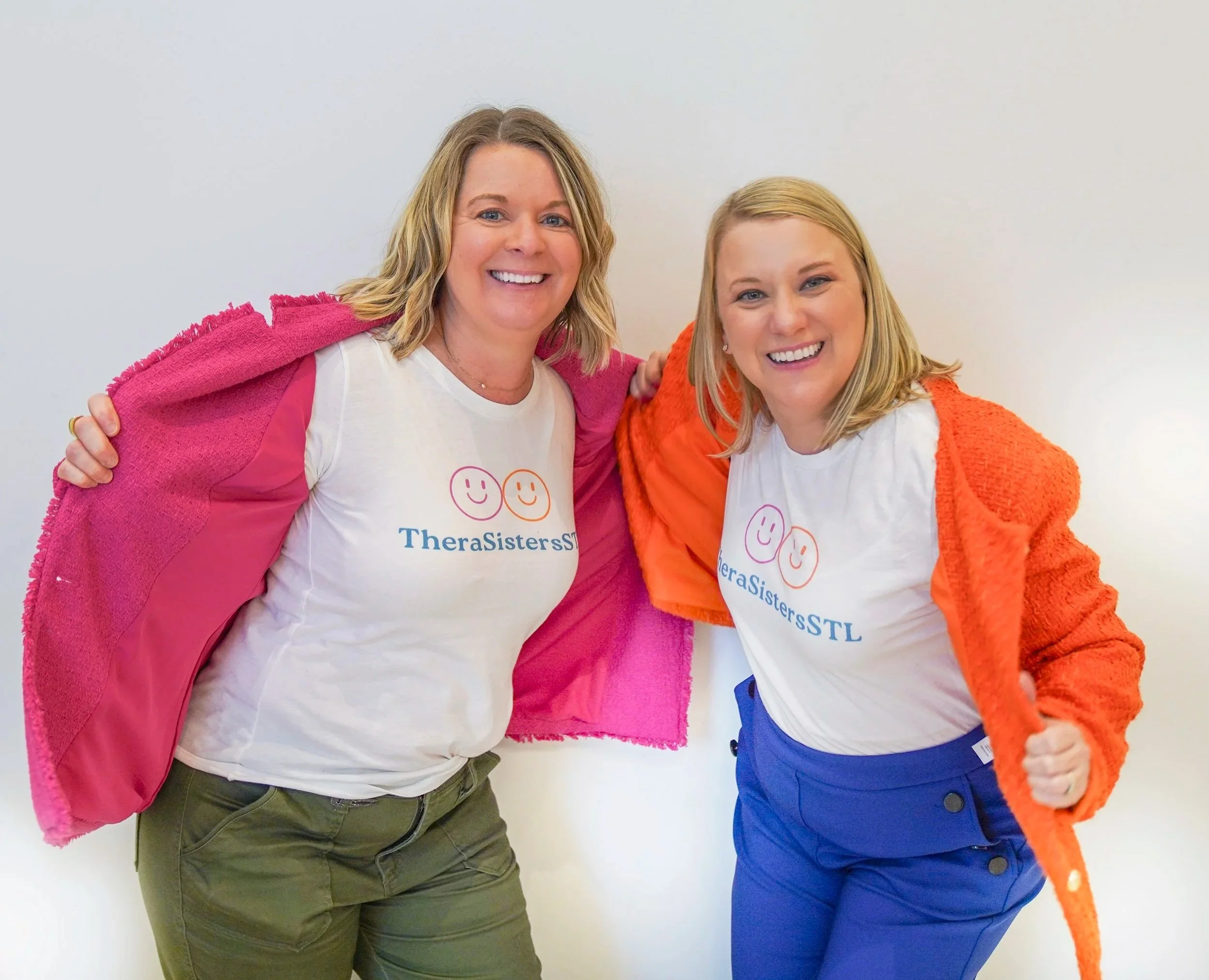 The TheraSistersStL, Amy and Katheryn smiling at the camera and showing their TheraSistersSTL t-shirts under their pink and orange coats