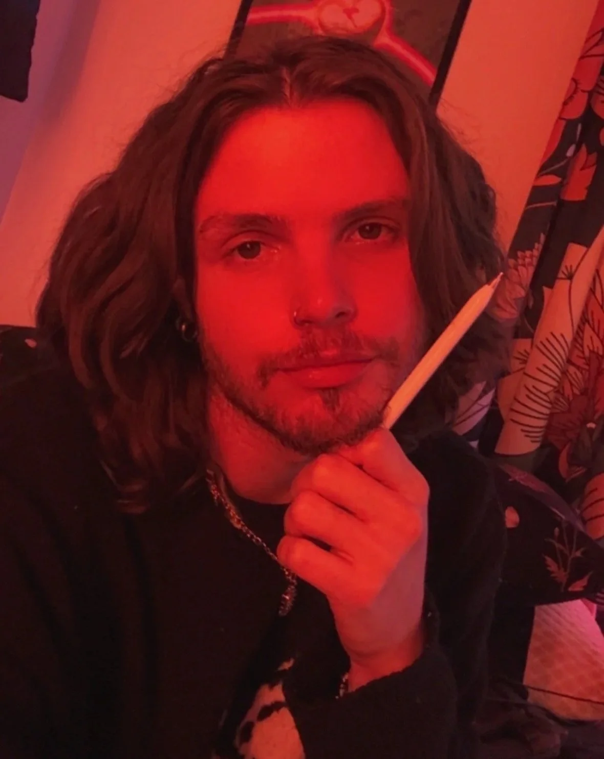 A person with long, wavy hair, a nose ring, and a small beard, holding an apple pencil close to his face while smiling slightly. The scene is bathed in red light and he is in a room with patterned curtains.