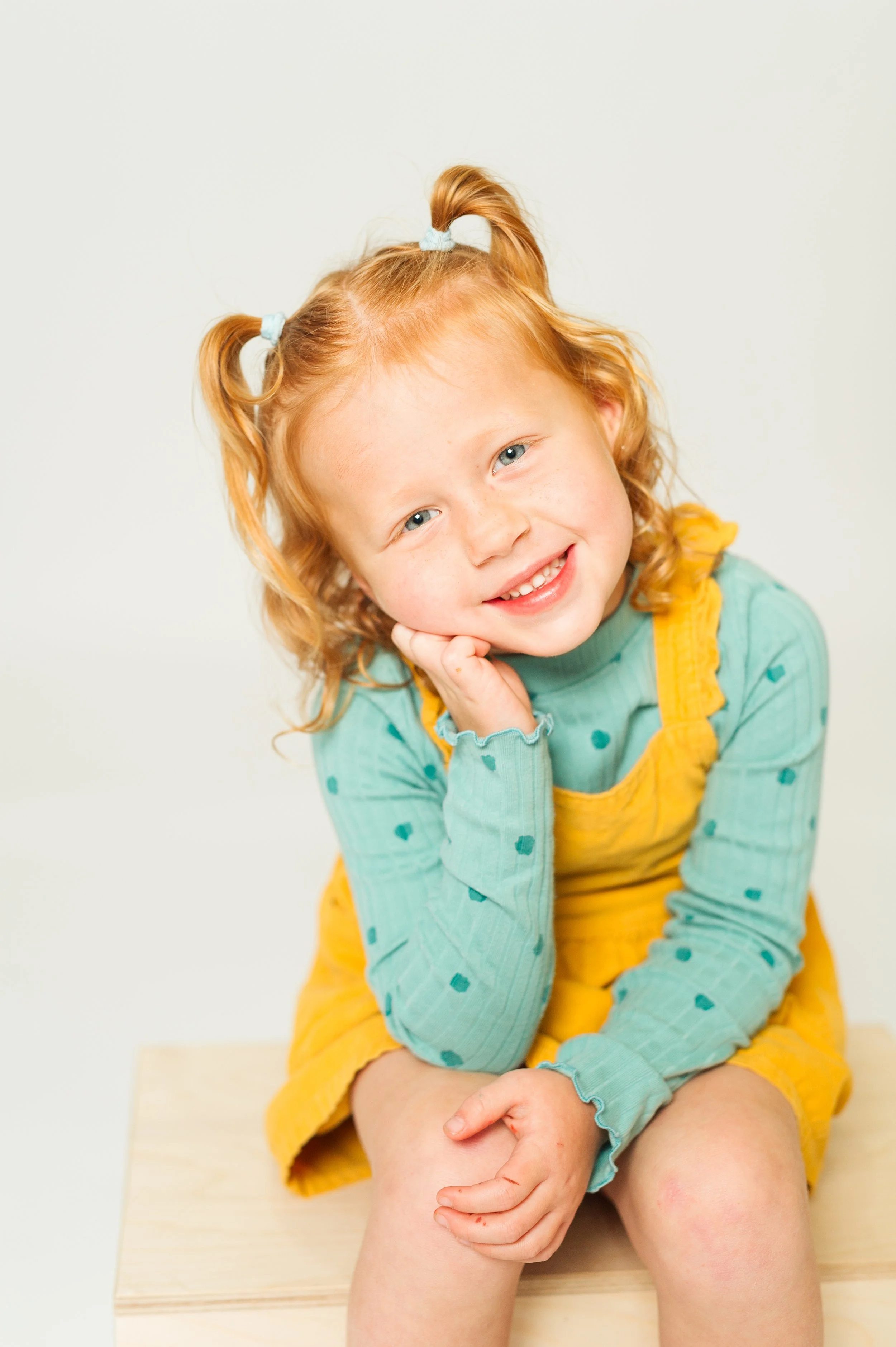 Smiling young child with red hair in pigtails wearing a turquoise shirt with polka dots and a yellow dress, sitting on a wooden box.