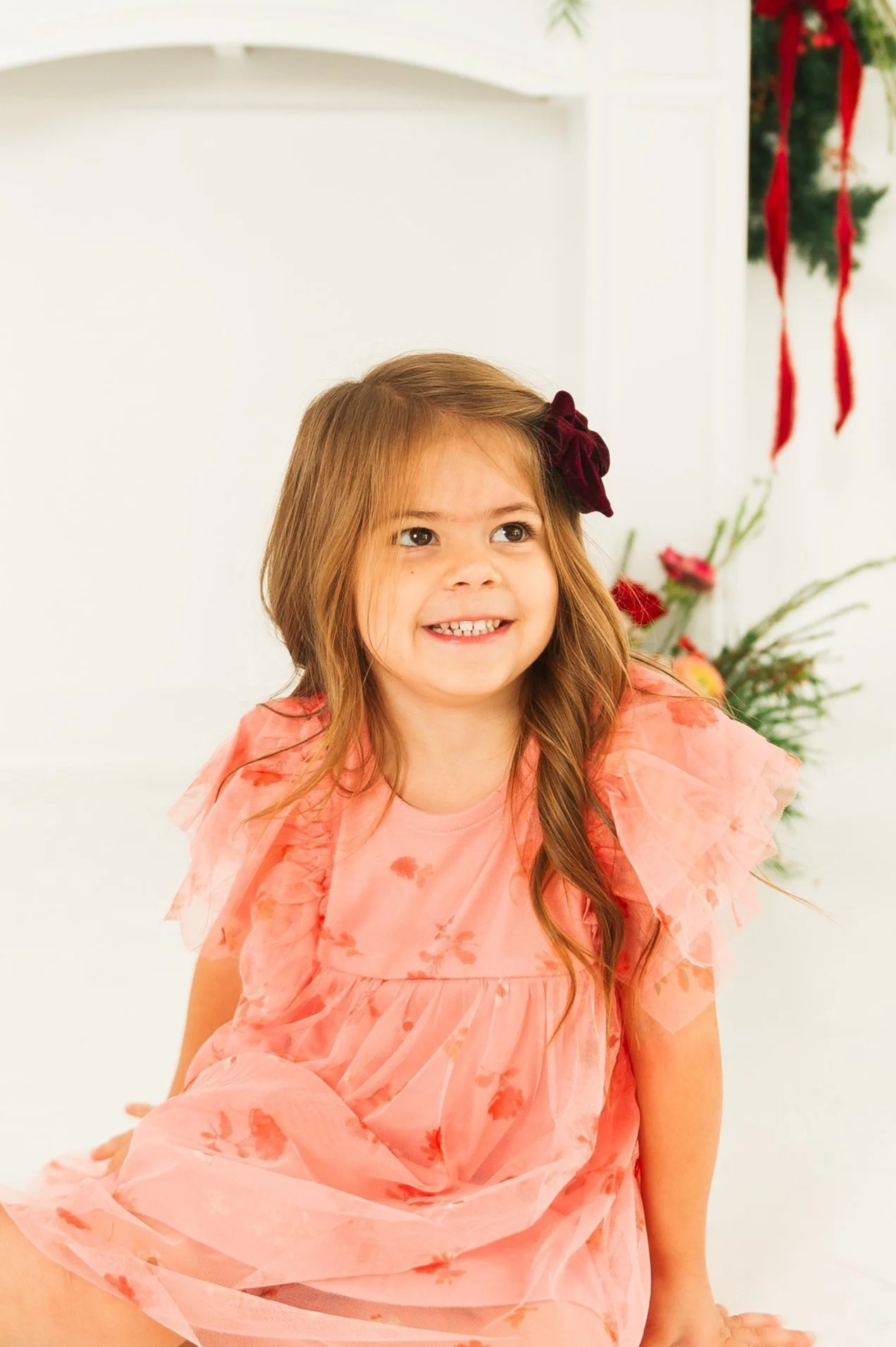 Young girl in a pink dress with floral patterns sitting indoors, with festive green and red decorations in the background.